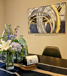 a table with flowers and wine bottles on it in front of a framed art piece