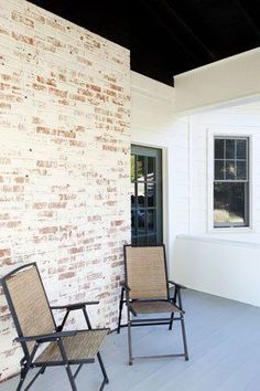 two chairs are sitting on the porch next to a brick wall and windows with white trim