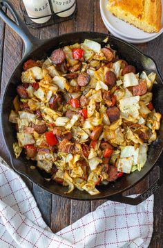 a skillet filled with sausage and vegetables on top of a wooden table next to a fork