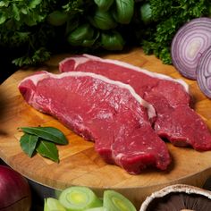 two raw meats on a wooden cutting board surrounded by vegetables and herbs, including onions