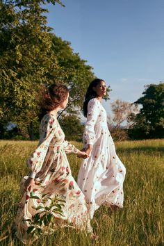 two women in dresses are walking through the tall grass with trees in the back ground