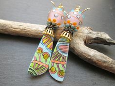 These earrings feature long abstract art ceramic charms by Sarreve in France. The charms are decorated with a whimsical pattern of shapes and lines in orange, yellow, black and blue against a pastel blue background.  They remind me a bit of alien life. Above the charms are long gold bead caps followed by lacey black brass bead caps.  Next are enamel and rhinestone flower bead caps in blue.  These support stunning matte lamp-work glass beads by Unventdeliberte in France. The lamp-work beads have Whimsical Earrings, Ceramic Charms, Artsy Earrings, Pastel Blue Background, Alien Life, Ceramic Earrings, Bleu Pastel, Colorful Ceramics, Ceramic Earring