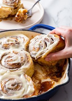 a person holding a piece of cinnamon roll in a blue casserole dish on a white table