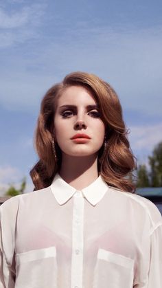 a woman with long hair wearing a white shirt and earrings, standing in front of a blue sky