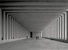 two people are walking down an empty hallway with columns on both sides and one person is standing in the middle