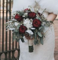 a bride holding a bouquet of red roses and white carnations in front of an iron gate