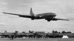 an airplane is flying low to the ground in front of some buildings and people standing around