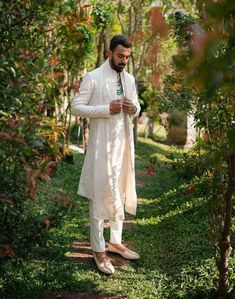 a man in a white suit and tie standing on the side of a tree lined path