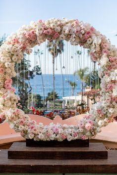 a circular floral arrangement on a wooden table in front of the ocean and palm trees