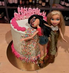 two dolls standing next to a birthday cake with sprinkles and candles on it