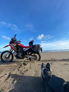 a motorcycle parked on top of a sandy beach next to the ocean with someone's feet in the sand