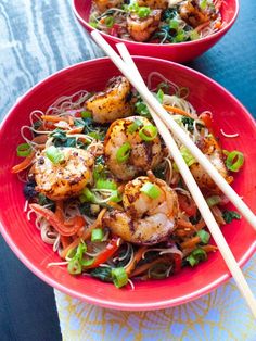 two red bowls filled with shrimp, noodles and veggies next to chopsticks