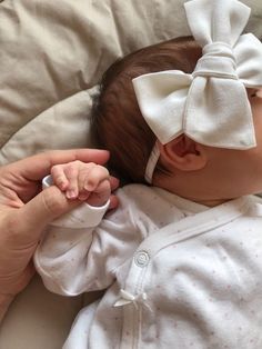 a baby with a big white bow on its head laying in a bed next to someone's hand