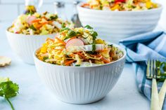 two white bowls filled with colorful salad on top of a blue table cloth next to silverware