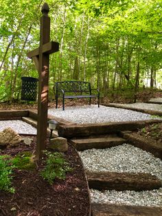 a wooden cross sitting in the middle of a forest filled with rocks and gravel next to a bench