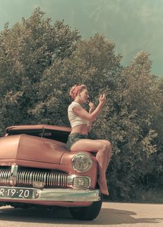 a woman is sitting on the hood of an old car with trees in the background