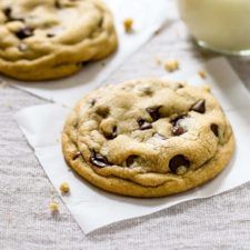 two chocolate chip cookies sitting on top of paper next to a glass filled with milk