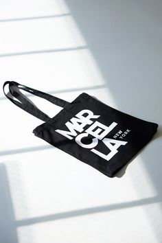 A canvas bucket tote bag in 100% European cotton, with a bottom panel for added capacity. The tote features an oversized MARCELLA NEW YORK graphic on one side, in white print on black canvas.[SPLIT] Our production team enjoys a living wage, a 40-hour work week with paid overtime, free health insurance and 20 days annual paid vacation. Black Rectangular Canvas Bag For Weekend, Casual Black Bags With Graphic Design, Large Capacity Black Cotton Canvas Bag, Cotton Tote Shoulder Bag With Logo Print, Black Canvas Weekend Bag, Large Capacity Cotton Bucket Canvas Bag, Black Canvas Bag For Weekend, Black Canvas Bag For The Weekend, Travel Cotton Shoulder Bag With Logo Print