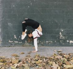 a woman doing a handstand in front of a brick wall with leaves on the ground