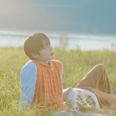 a young man sitting on top of a lush green field next to a lake and grass covered ground