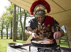 a man dressed in roman armor cooking food on an outdoor grill with trees in the background