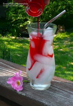 a glass filled with liquid and ice sitting on top of a wooden table next to a pink flower