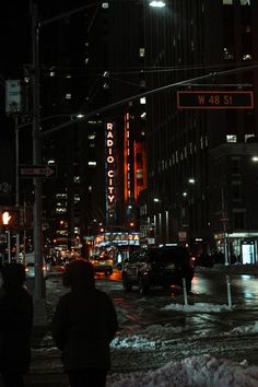 people are walking in the snow on a city street at night with tall buildings behind them