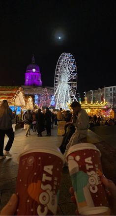 people walking around an amusement park at night with ferris wheel in the background and lights on