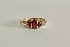 This is a gorgeous Edwardian Era 14k Yellow Gold Garnet Ring with a three genuine garnets and four white sapphire gemstone setting, the middle garnet is oval faceted cut the two side ones are faceted round cut, with four rose cut white sapphires one on each corner. A stunning piece of fine early 1900s era jewelry, featuring January's birthstone! ERA - Circa 1900s / Edwardian METAL / MATERIAL - 14k yellow gold, 3 genuine garnets (approx. 0.77 CTW), 4 white sapphires (approx. 0.02 CTW) MARKINGS / Gold And Garnet Jewelry, Art Deco Yellow Gold Three-stone Ring, Antique 14k Gold Three-stone Diamond Ring, Antique Three-stone Ruby Wedding Ring, Heirloom Ruby Ring With Three Stones In Round Cut, Heirloom Ruby Three-stone Ring, Heirloom Ruby Ring With Three Stones, Heirloom Ruby Three Stone Ring, Elegant Red Three-stone Birthstone Ring