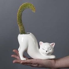 a hand holding a white ceramic cat with a green plant in it's mouth