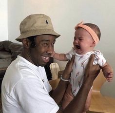 a man holding a baby in his right hand and smiling at the camera while wearing a hat