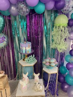 a table topped with cakes and balloons in front of purple, blue and green decorations