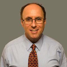 a man wearing glasses and a tie smiling at the camera with his hands in his pockets