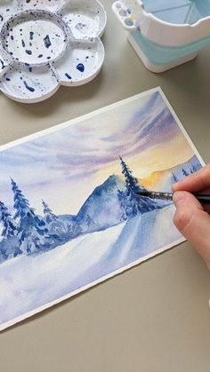 someone is painting a snowy mountain scene with watercolors on the table next to some dishes