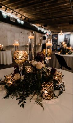 candles and flowers on a white table cloth with greenery in the center, surrounded by glass vases