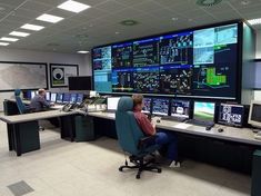 two men are sitting in front of multiple monitors on the wall, and one man is looking at them