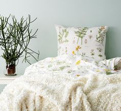 a white bed topped with pillows and blankets next to a potted plant on top of a table