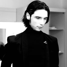 a man with long hair wearing a black suit and turtle - neck sweater looking at the camera