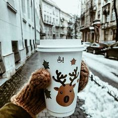 someone holding up a coffee cup in the middle of a snowy street with buildings and cars