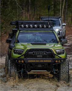 the front end of a green toyota truck driving through muddy terrain with two other trucks in the background