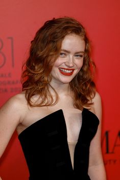 a woman with red hair wearing a black dress and smiling at the camera while standing in front of a red wall