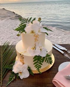 a wedding cake with white flowers and greenery sits on a table next to the ocean