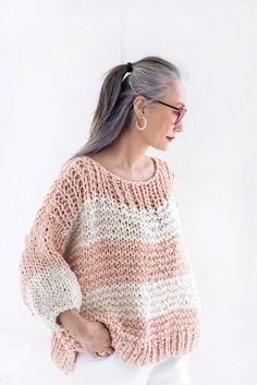 an older woman with grey hair wearing a pink and white striped sweater, standing in front of a white wall