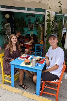 two people sitting at a table with plates of food in front of them on the sidewalk