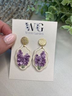 a pair of earrings with purple flowers in them on a white card next to a plant