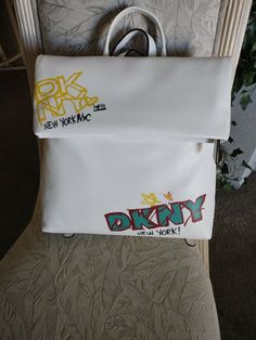 AUTHENTIC “ DKNY” WOMEN’S TILLY WHITE SIGNATURE MD FOLDOVER LEATHER BACKPACK! BRAND NEW/W/TAGS! NEVER USED IN PERFECT CONDITION! STYLE # R12Q350 COLOR: WHITE ONE SIZE M.S.R.P. 168.00 PLUS TAX! FOLDOVER  ZIPPER OPEN/CLOSE W/MAGNETIC SNAP IN CENTER ZIPPER HAS LEATHER PULL TAB JUST UNDER THE FOLDOVER IS A LARGE ZIPPER POCKET THAT  MEASURES: 9” (L) 10.5” (H) ALSO W/WHITE LEATHER PULL TAB SILVER TONE HARDWARE  FRONT OF THE BACKPACK HAS DESIGNER NAME IN YELLOW AND SAYS” LOVE NEW YORK, N Trendy White Backpack, Trendy White Leather Backpack, Trendy Logo Backpack Bag, White Backpack For Shopping, Trendy Logo Backpack, Trendy White Bags For Streetwear, Trendy White Streetwear Bags, Designer Name, R P