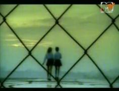 two people standing on the beach looking out at the ocean from behind a chain link fence