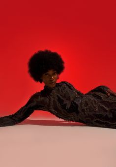 a woman with an afro laying on the floor in front of a red background wearing a black dress