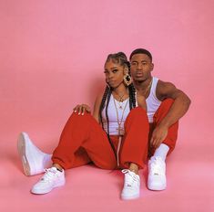 a man and woman sitting next to each other on a pink background wearing white sneakers