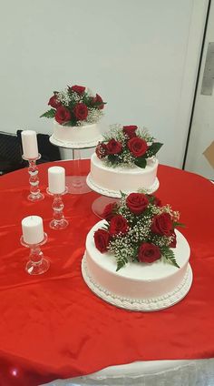 three tiered white wedding cake with red roses on top, surrounded by wine glasses and candles